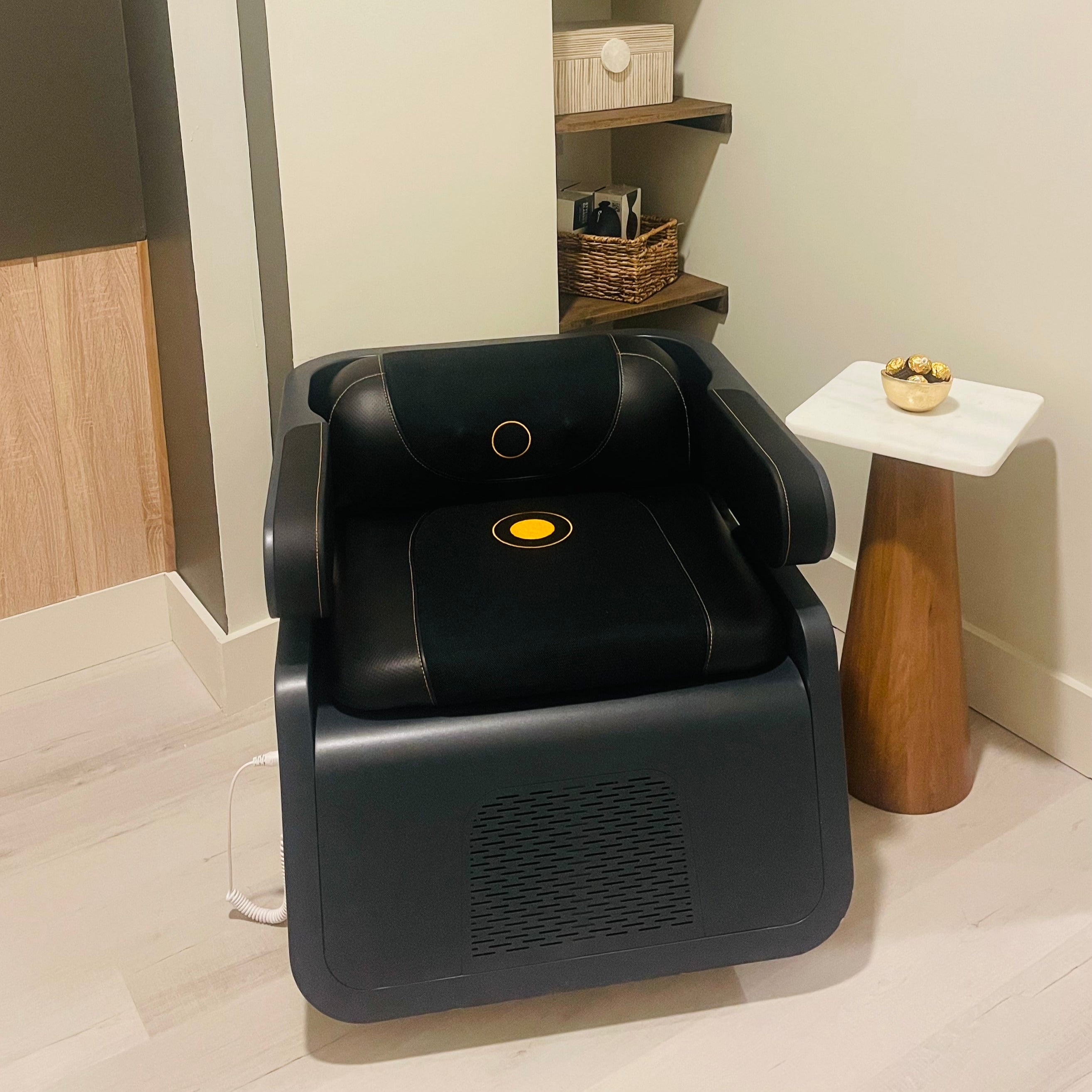 Black massage chair in a room with shelves and a small table.